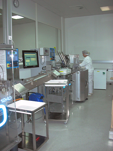 Pomieszczenie pakowania farmaceutycznego pokazuje, że trasy operatora, komponentów, produktu i ruchomego wyposażenia wyznacza rzeczywiste ustawienie linii, a nie tylko strzałki na diagramie. Źródło: [Wikimedia Commons, „Pharma-production.jpg”](https://commons.wikimedia.org/wiki/File:Pharma-production.jpg), Peter Robert Binter, domena publiczna.